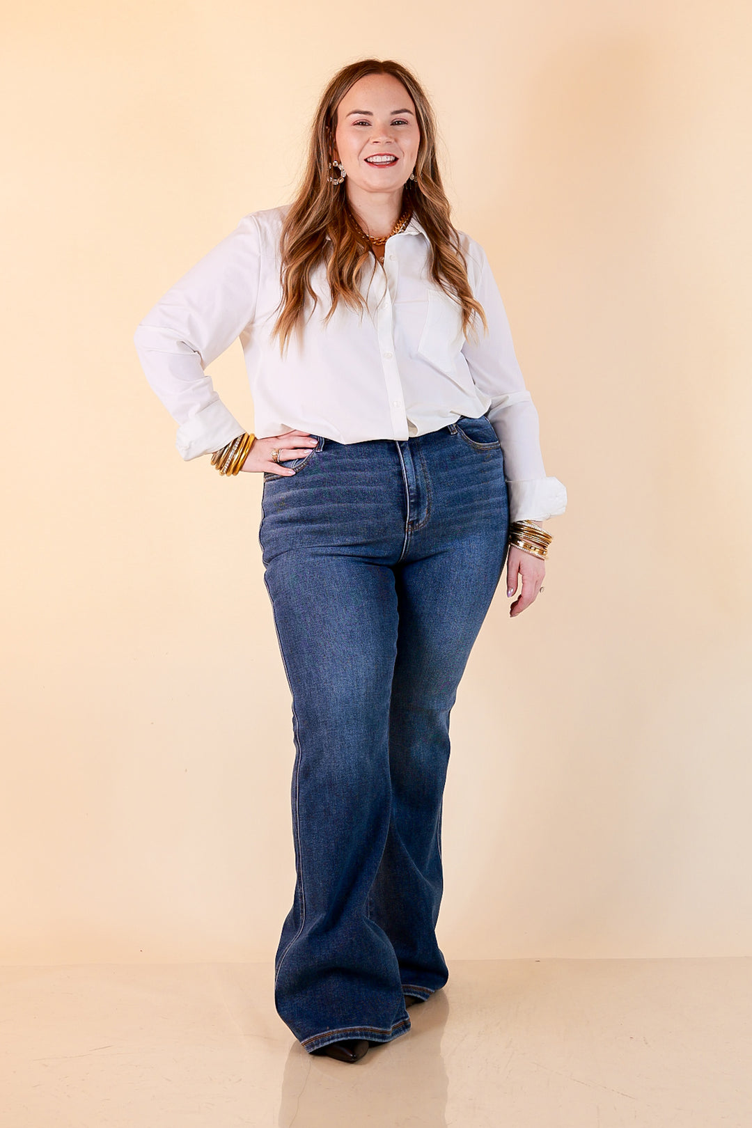 A woman in a white button-up shirt and medium wash Judy Blue flare jeans with gold bangles and a gold chain necklace