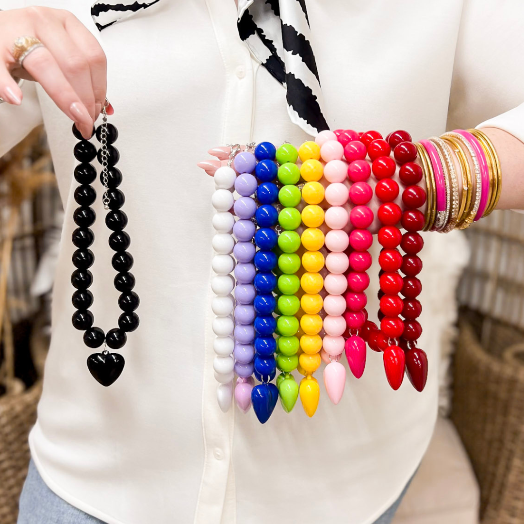 A woman in a white blouse holds up seven strands of spike bead necklaces in black, lavender, blue, green, yellow, hot pink and red, with stacked gold bangles on her wrist.