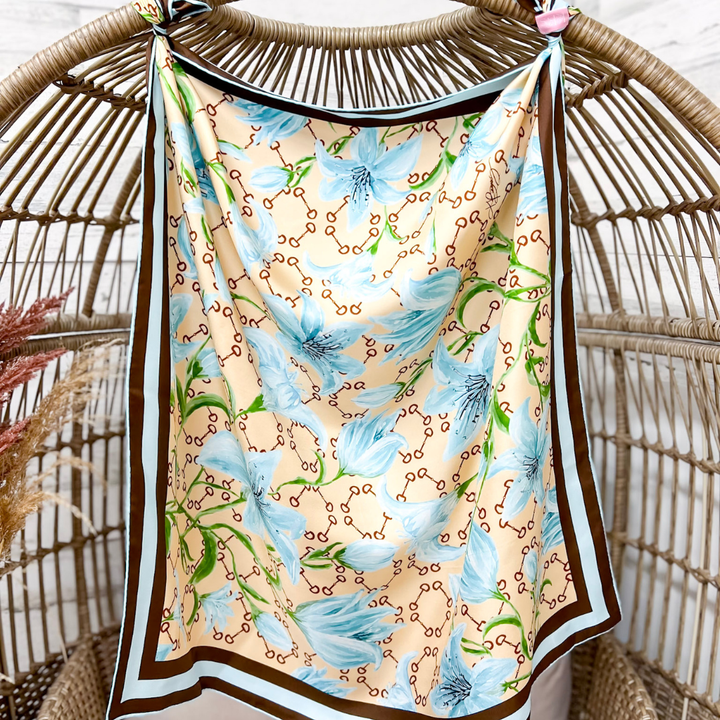 A square silk scarf with a beige background, decorated with light blue feathers, green vines, and brown horsebit chain links, hanging from a woven rattan stand.