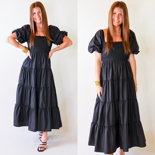 Model is wearing a long black dress featuring puffed balloon sleeves, smocked bodice, and tiered skirt.