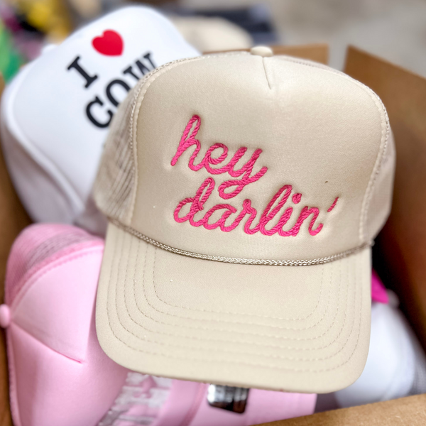 Photo pictures a cream colored foam trucker hat featuring pink cursive embroidery of the words "Hey darlin'"