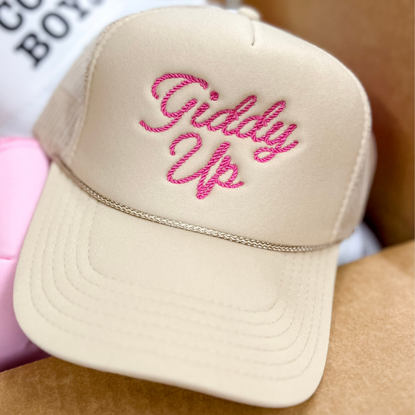 Photo features a tan trucker hat with pink cursive embroidery of the words giddy up.