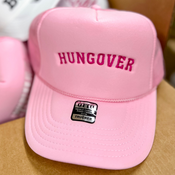 Photo pictures a light pink trucker hat featuring a hot pink embroidery of the word hungover.