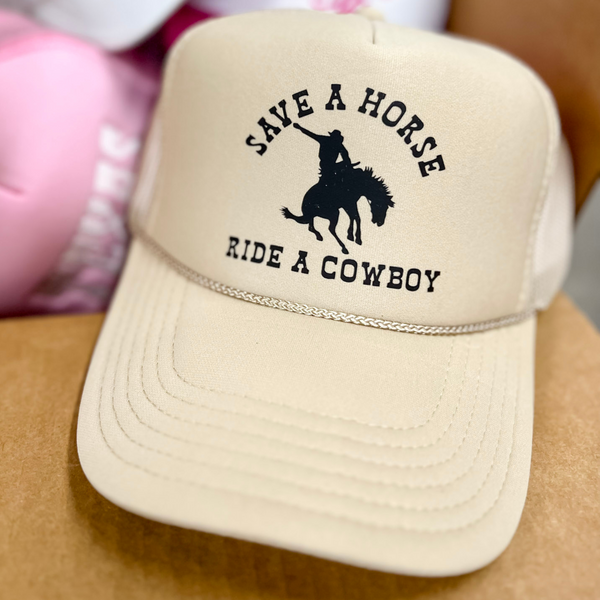 Photo features a tan trucker hat with the words "Save a Horse Ride a Cowboy" in black on the front. Also has a black graphic of a cowboy riding a horse