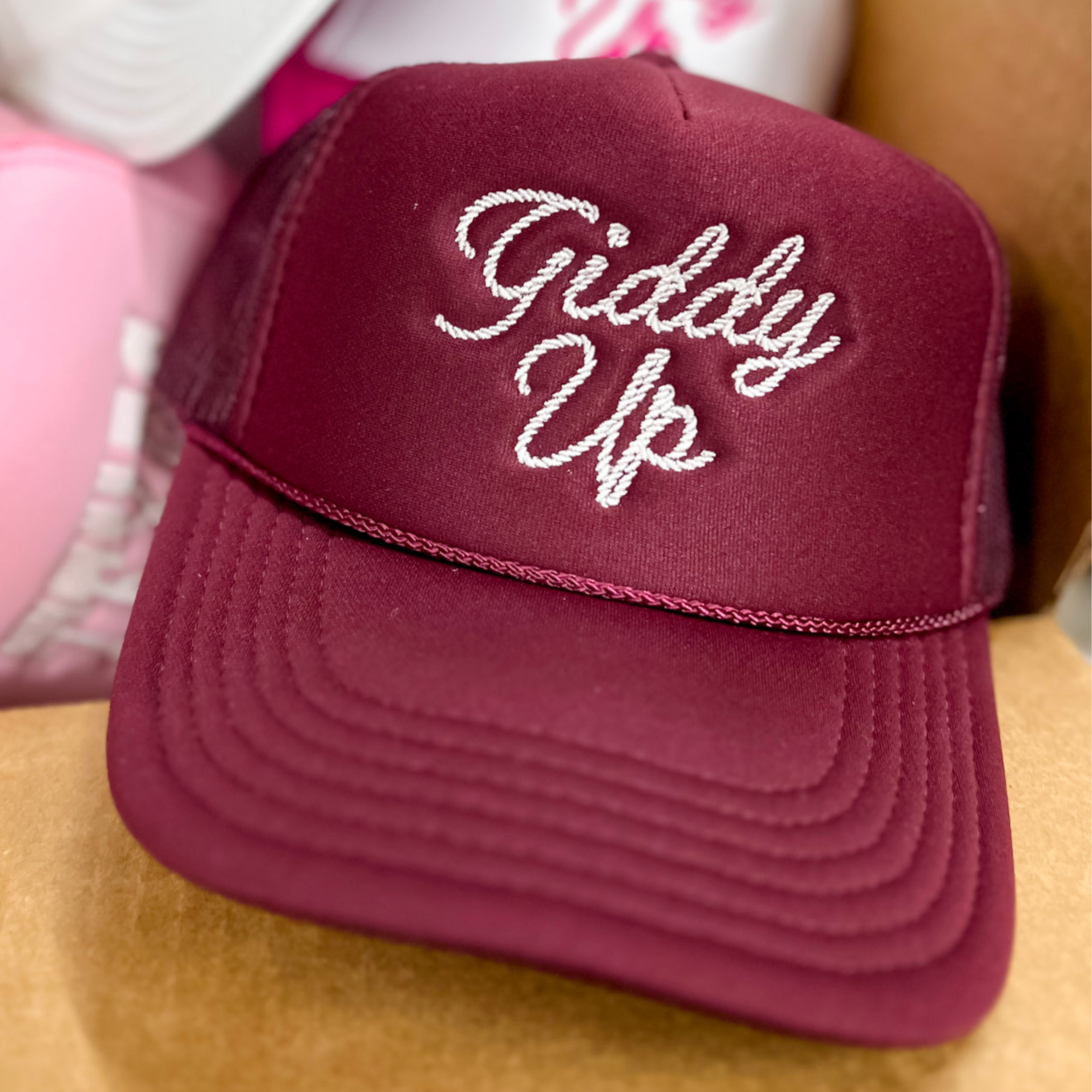 Photo features a maroon trucker hat with white cursive embroidery of the words giddy up on the front.