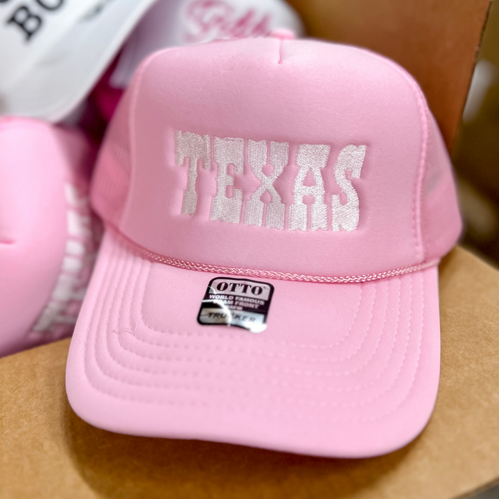 Photo features a light pink trucker hat with white bold embroidery of "Texas" on the front.