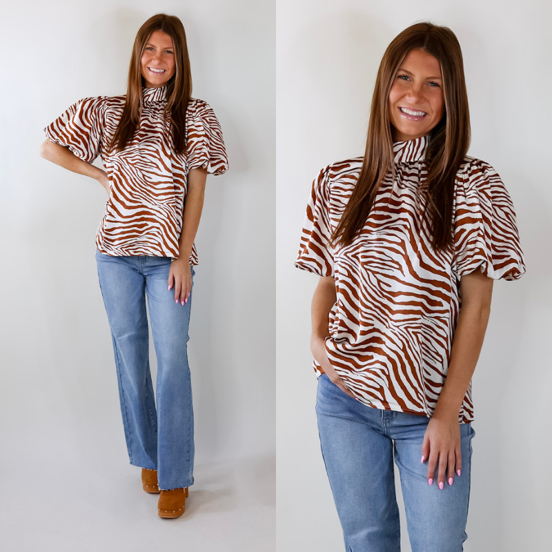 Model is wearing a ginger and white zebra print top with balloon sleeves and a high neckline that ties in the back. Model has paired the top with light wash jeans, and camel brown booties. 