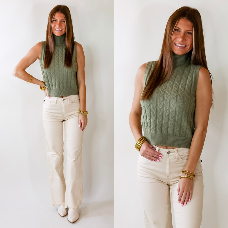 Model is wearing a sleeveless sage green cropped sweater with a high neckline. Model has paired the sweater with white jeans, white booties, and gold tone jewelry. 