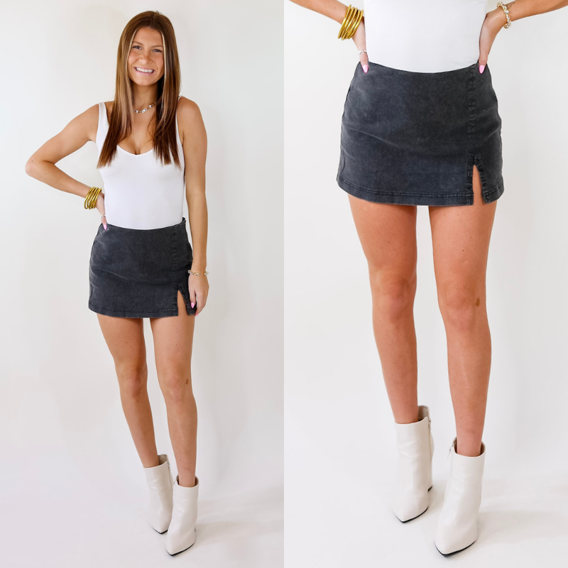 Model is wearing a skort with a side cut, side zipper, and a black denim wash. Model has paired the skort with a white bodysuit, white booties, and gold tone jewelry. 