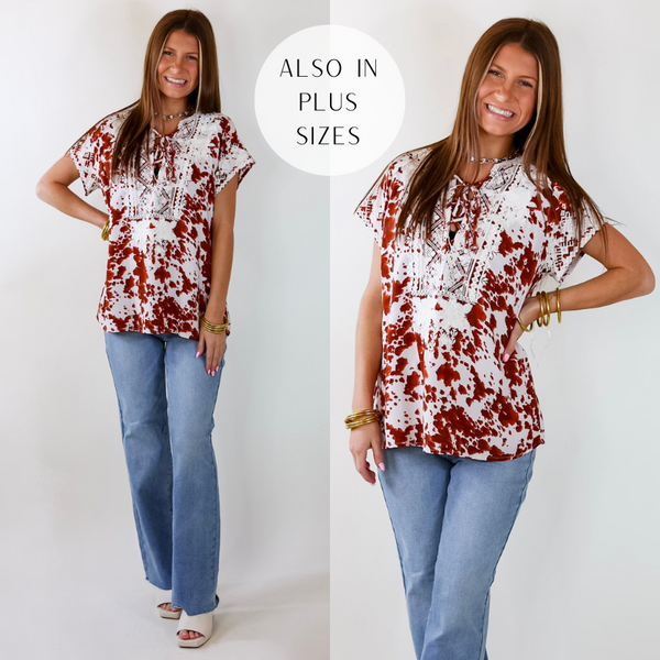 Model is wearing a rust orange spotted top with white geometric and floral embroidery and a notched neckline that ties in the front. Model has paired the top with straight cut jeans, ivory heels, and gold tone jewelry. 