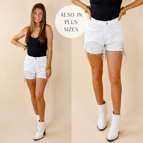 Model is wearing a pair of white corduroy shorts with crystal fringe on both legs. Model has paired these shorts with a black bodysuit, white booties, and gold tone jewelry.
