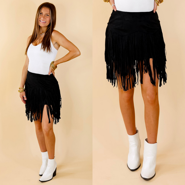 Model is wearing a black skirt with fringe in a zig zag pattern. Model has paired the skirt with a white bodysuit, white booties, and gold tone jewelry. 