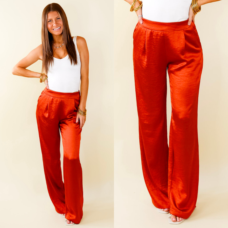 Model is wearing a pair of rust orange silky pants. Model has paired the pants with a white bodysuit, white heels, and gold tone jewelry. 