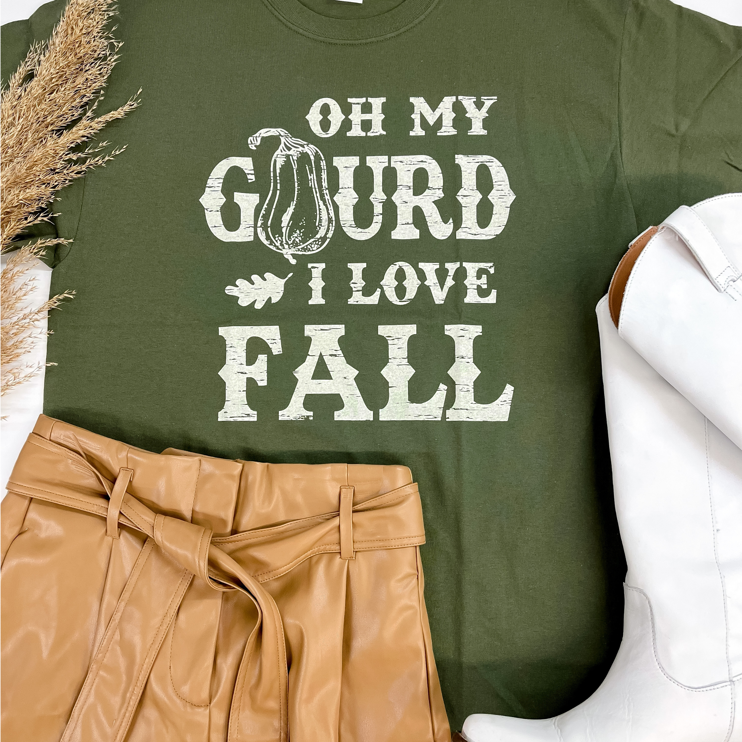 A folded tee shirt that is moss green. The t-shirt has white lettering that says "Oh my Gourd I love fall". Pictured on a white background with tan faux leather shorts and white boots.