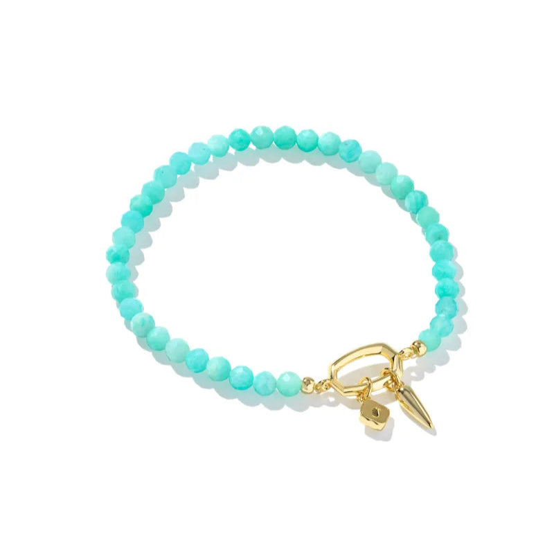 A solid white background with a teal beaded bracelet by kendra scott with a gold toggle closure.