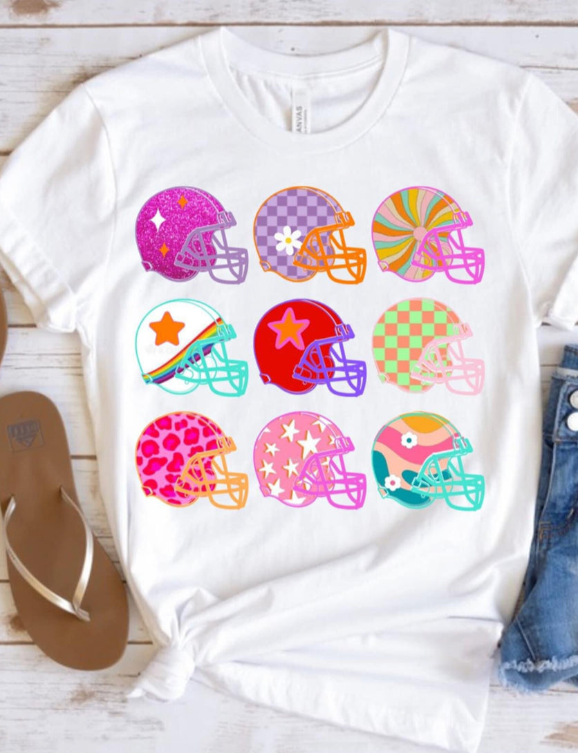 White graphic tee featuring nine colorful football helmets with floral, checkered, leopard, swirl, and retro-inspired patterns for a fun game day look.