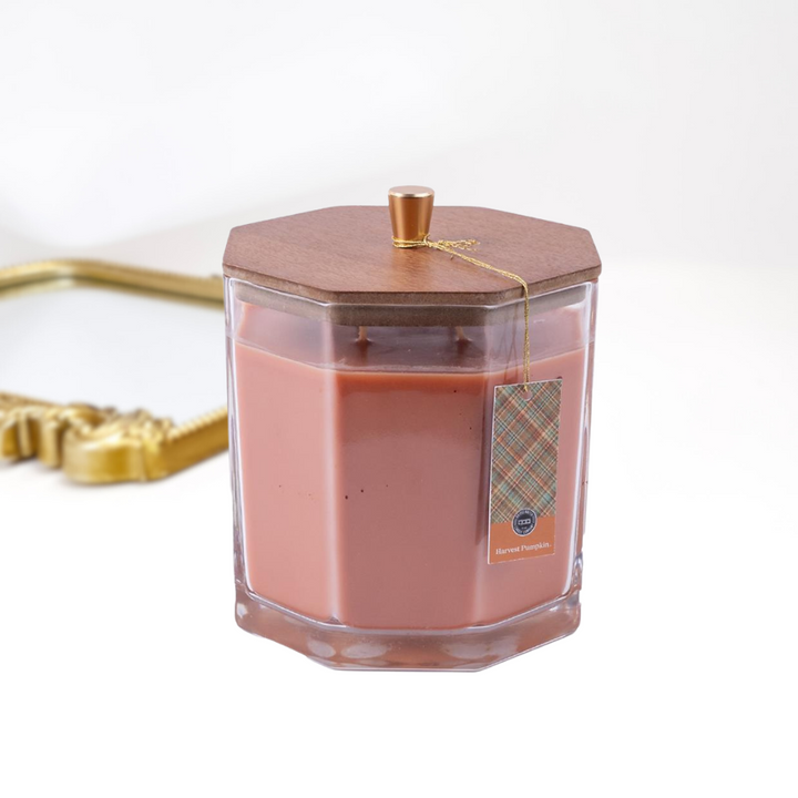 A solid white background with a gold mirror and a large candle in the front, The glass candle has a wooden with with a gold handle.