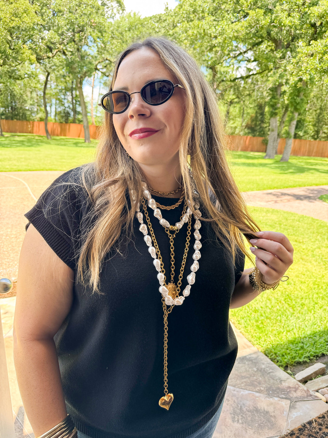 Woman wearing sunglasses and a long necklace outdoors with greenery in the background