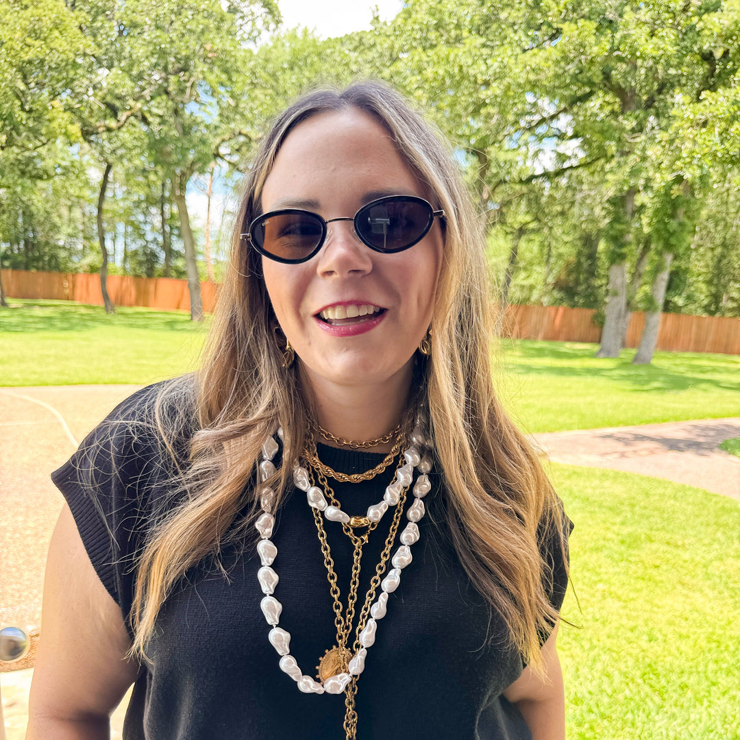 Woman wearing sunglasses and layered necklaces outdoors with greenery in the background