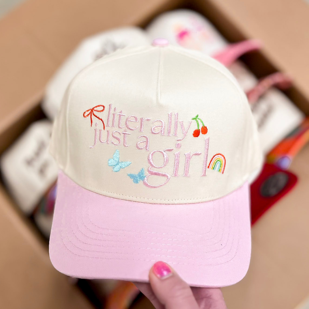 Pink and white cap with colorful text and graphics held by a hand in front of a cardboard box.