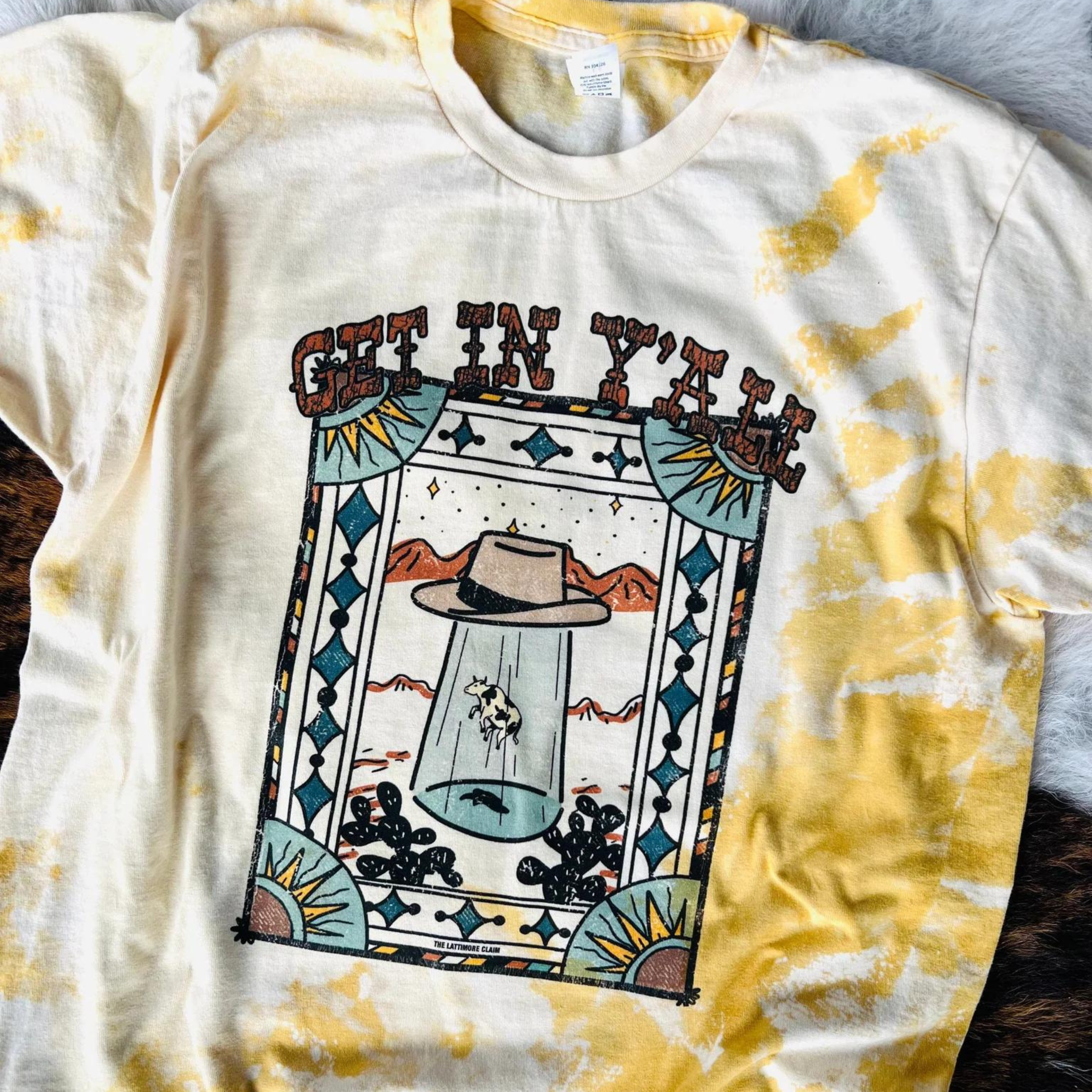 An orange short sleeve crewneck tee with a bleach splatter pattern. Featuring a graphic of a cow being abducted by a fedora in a desert. The graphic is surrounded by an artistic border of alternating colors, suns in the corners, and blue diamonds. The text "Get in yall" can be found right above the graphic in a western font. The item is pictured on a tiger print background 