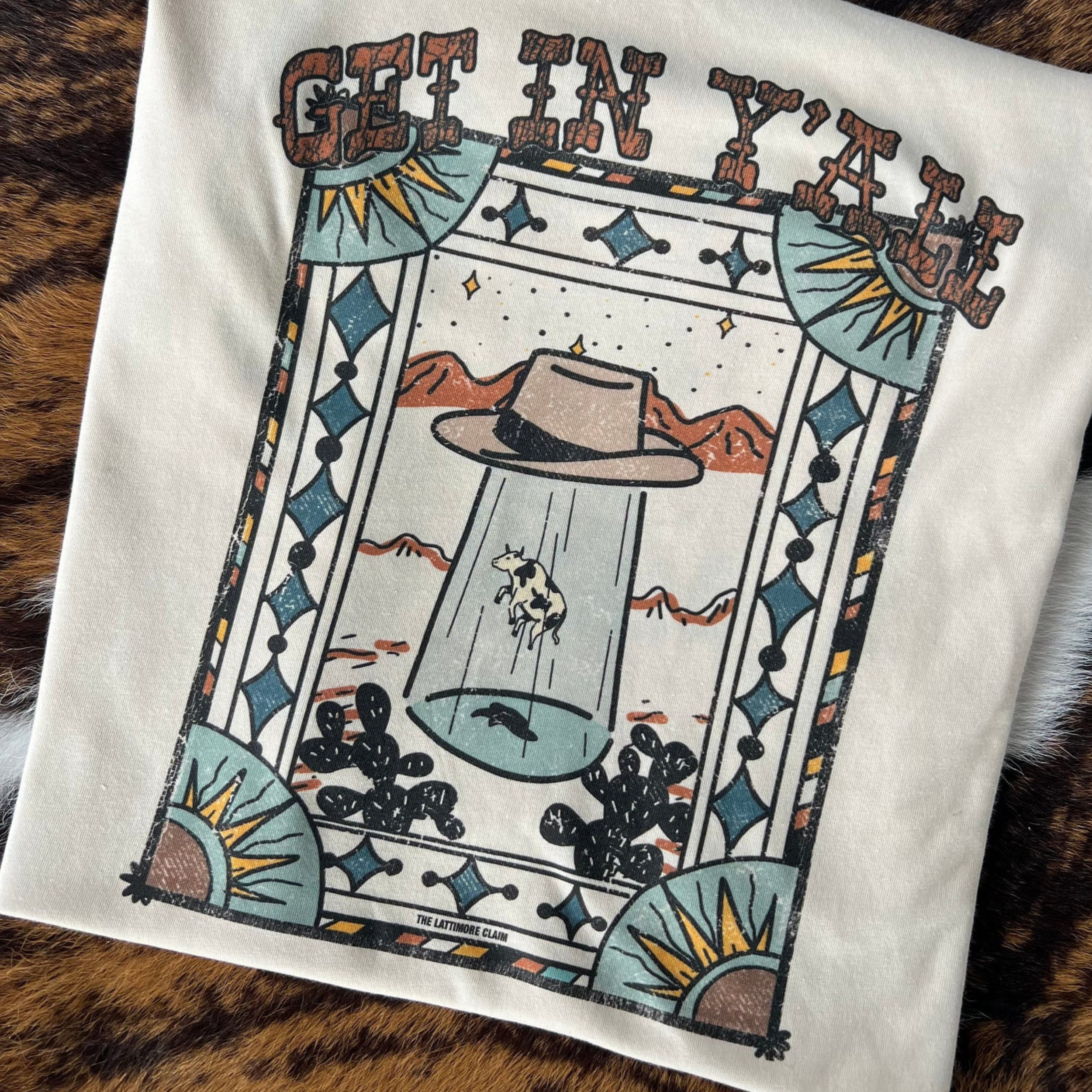 A white short sleeve crewneck tee with a graphic of a cow being abducted by a fedora in a desert. The graphic is surrounded by an artistic border of alternating colors, suns in the corners, and blue diamonds. The text "Get in yall" can be found right above the graphic in a western font. The item is pictured on a tiger print background