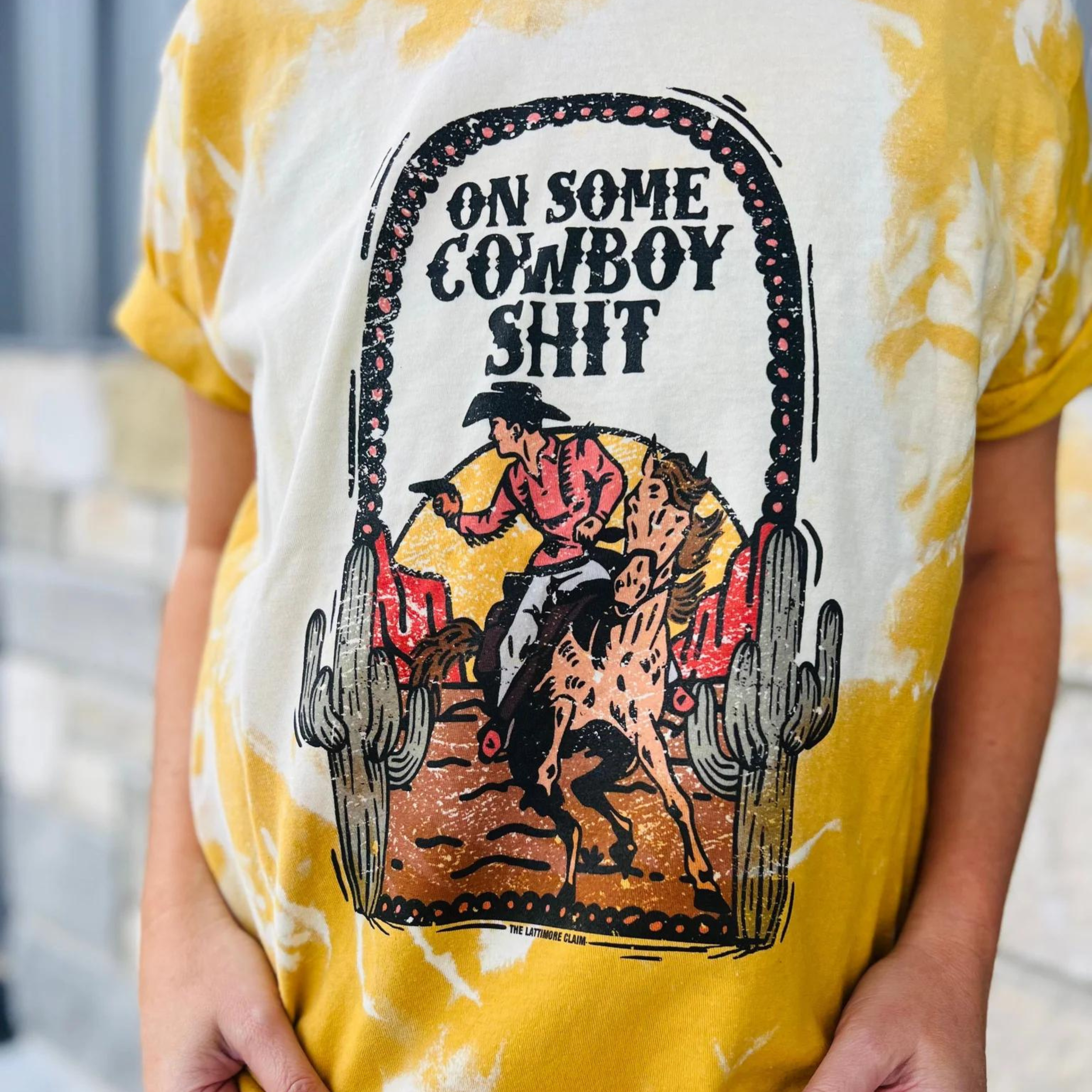 An orange with a bleach splatter pattern short sleeve crewneck tee with a graphic of a cowboy and his horse in the desert. The text "On some cowboy sh*t" is centered above the graphic. The item is pictured on a heather grey background
