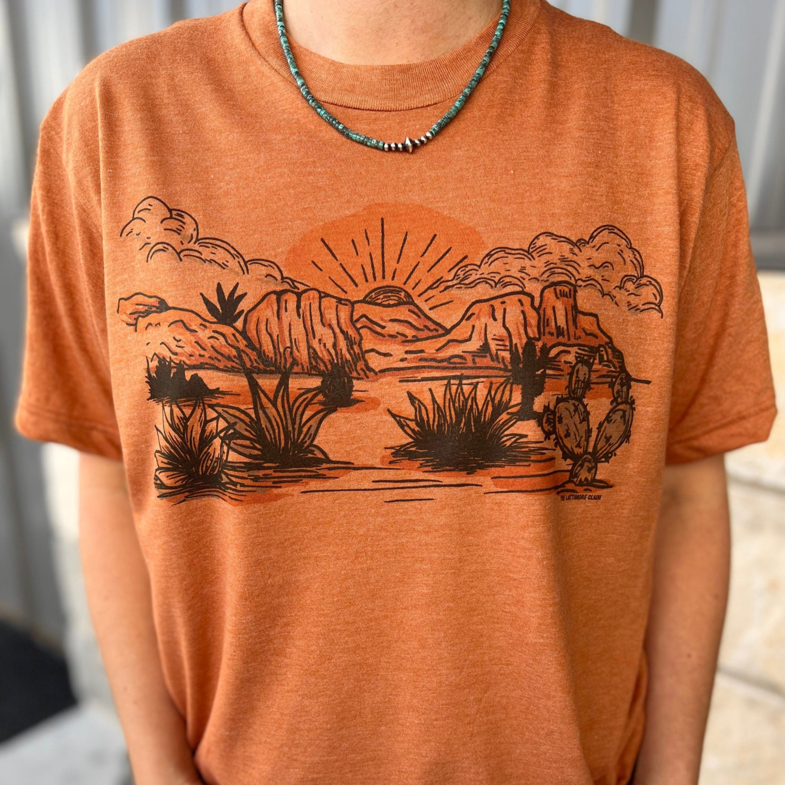 An orange short sleeve crewneck tee with a graphic in the center featuring cacti, desert flora, cliffs, and a sunset. Item is pictured on a multicolor background with white, grey, and black
