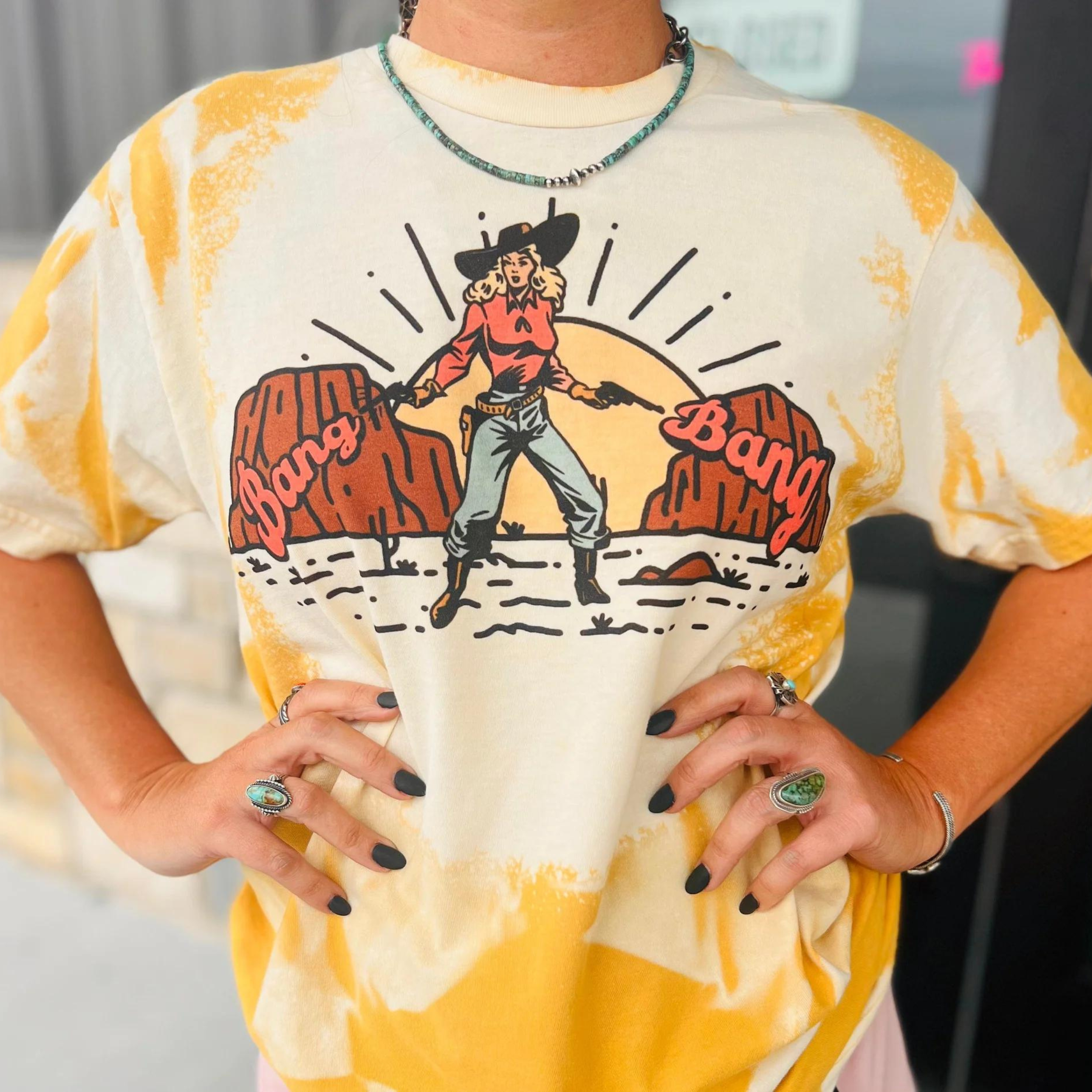 A mustard yellow tee with a bleached design. Featuring a cowgirl in a desert sunset wearing a hat, red shirt, blue jeans, and boots. The words "Bang Bang" are seen on either side of the cowgirl. Item is pictured on a casual background with hints of grey, white, and black.  
