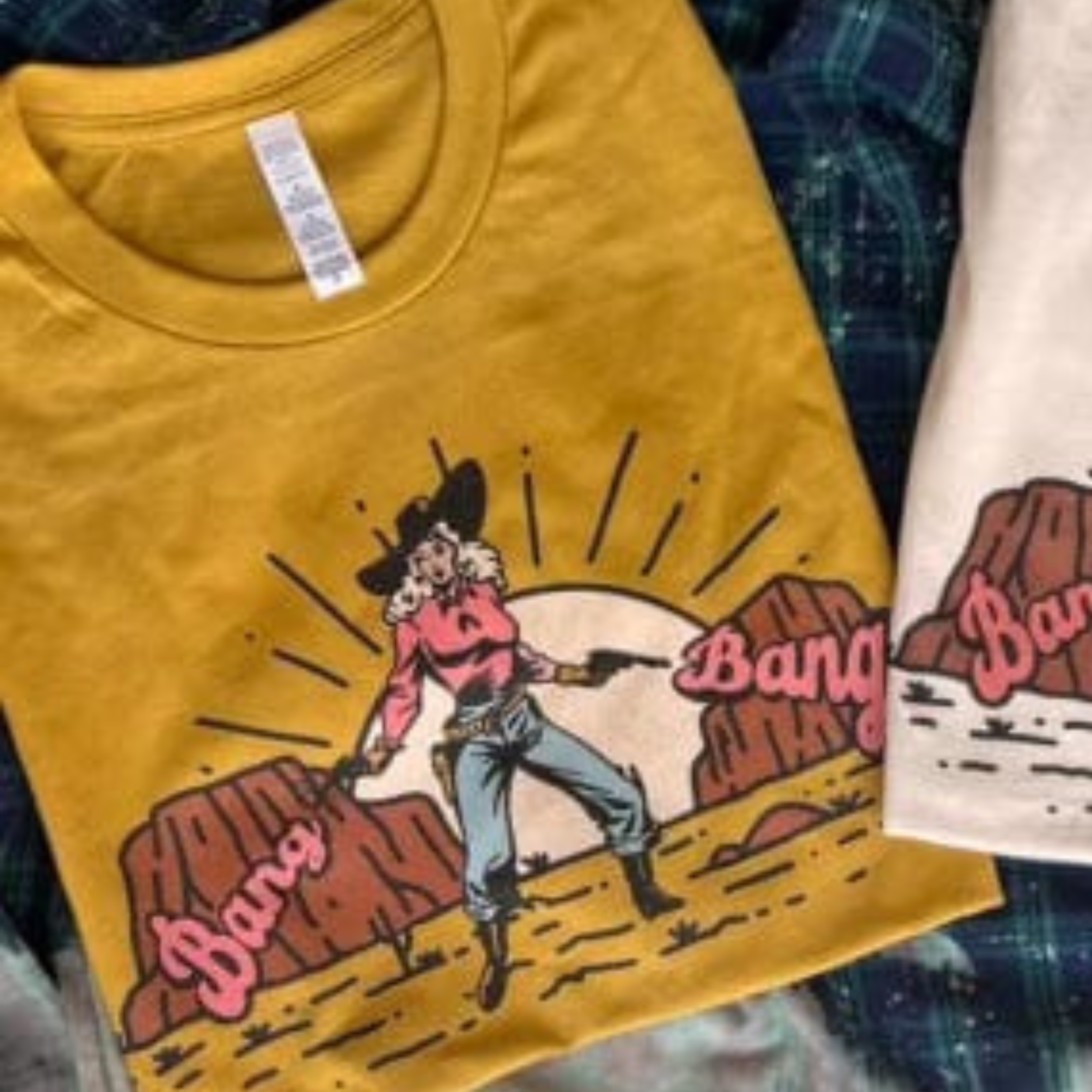 A mustard colored crewneck tee featuring a cowgirl in the desert at sunset wearing a black hat, red shirt, blue jeans, and boots. The words "Bang Bang" can be found on either side of the cowgirl. Item is pictured on a plaid background.