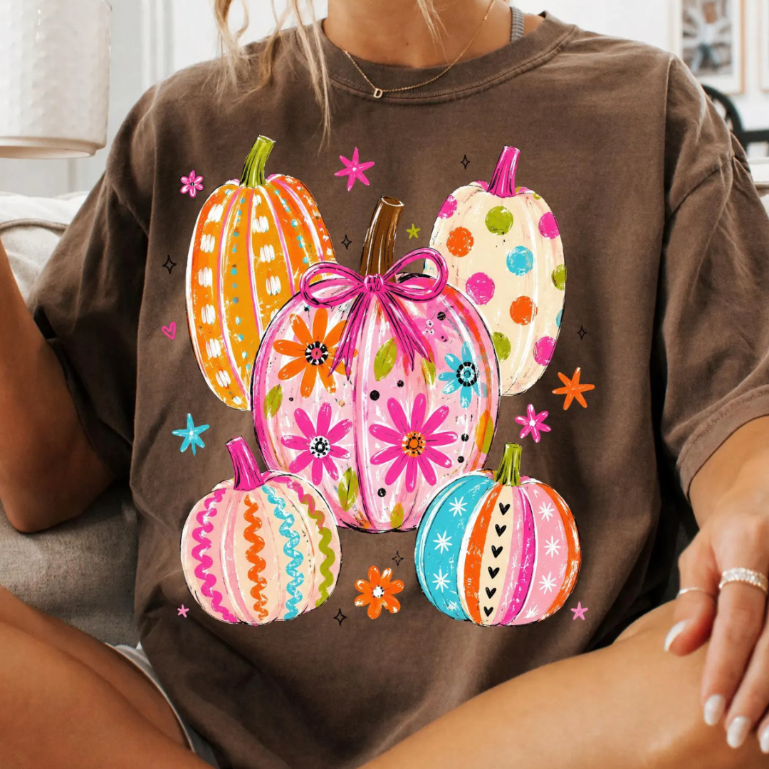 A person wearing a brown short-sleeve T-shirt with a colorful graphic of five decorated pumpkins and flowers on the front, photographed indoors.