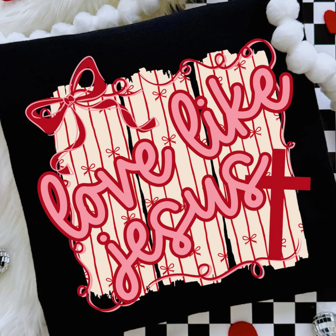 Black graphic tee featuring a pink and red Love Like Jesus design with a cream bow-print background, statement ribbon bow, and red cross.