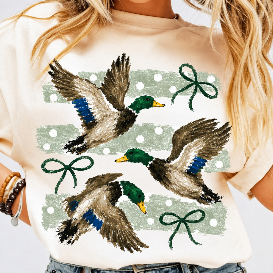 Ivory graphic tee or sweatshirt featuring three flying mallard ducks with sage green polka-dot brushstrokes and dark green bow accents.