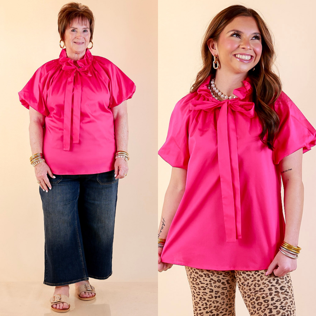 Models are wearing Short Puff Sleeve Tie Neck Blouse with Gathered Neckline in Fuchsia Pink with Jeans against a beige backdrop. 