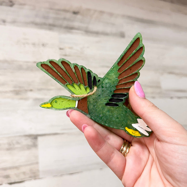 A hand holding a car freshie that is shaped like a duck, with a wooden background.