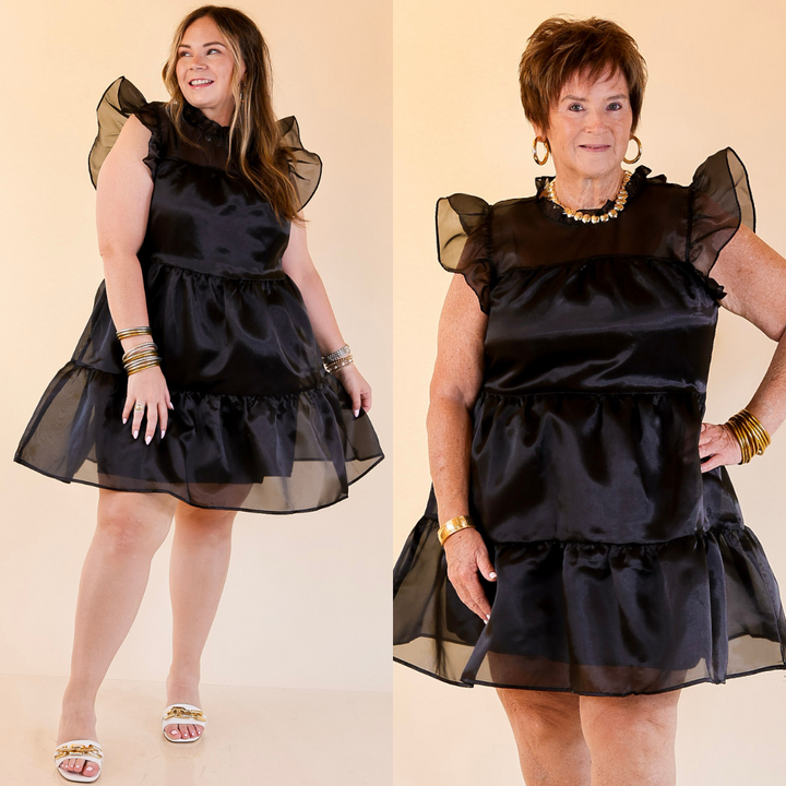 Two women wearing a black dress with sheer sleeves against a beige background