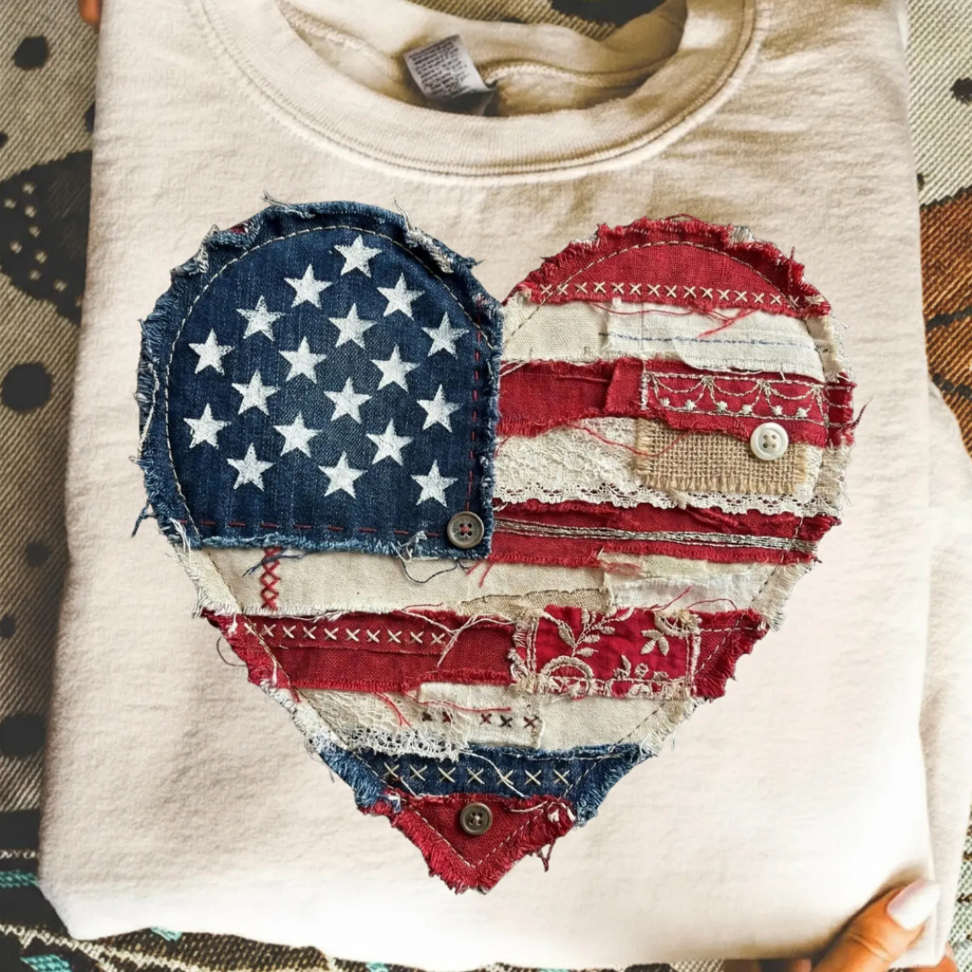 Natural beige graphic tee featuring a patchwork American flag heart with distressed denim, lace, button, and vintage patriotic details.