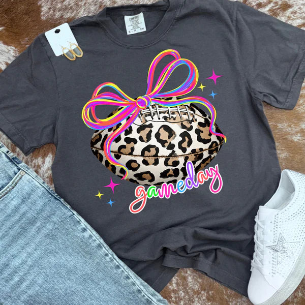 Gray t-shirt with leopard print football and rainbow bow graphic, paired with jeans and sneakers on a wooden surface.