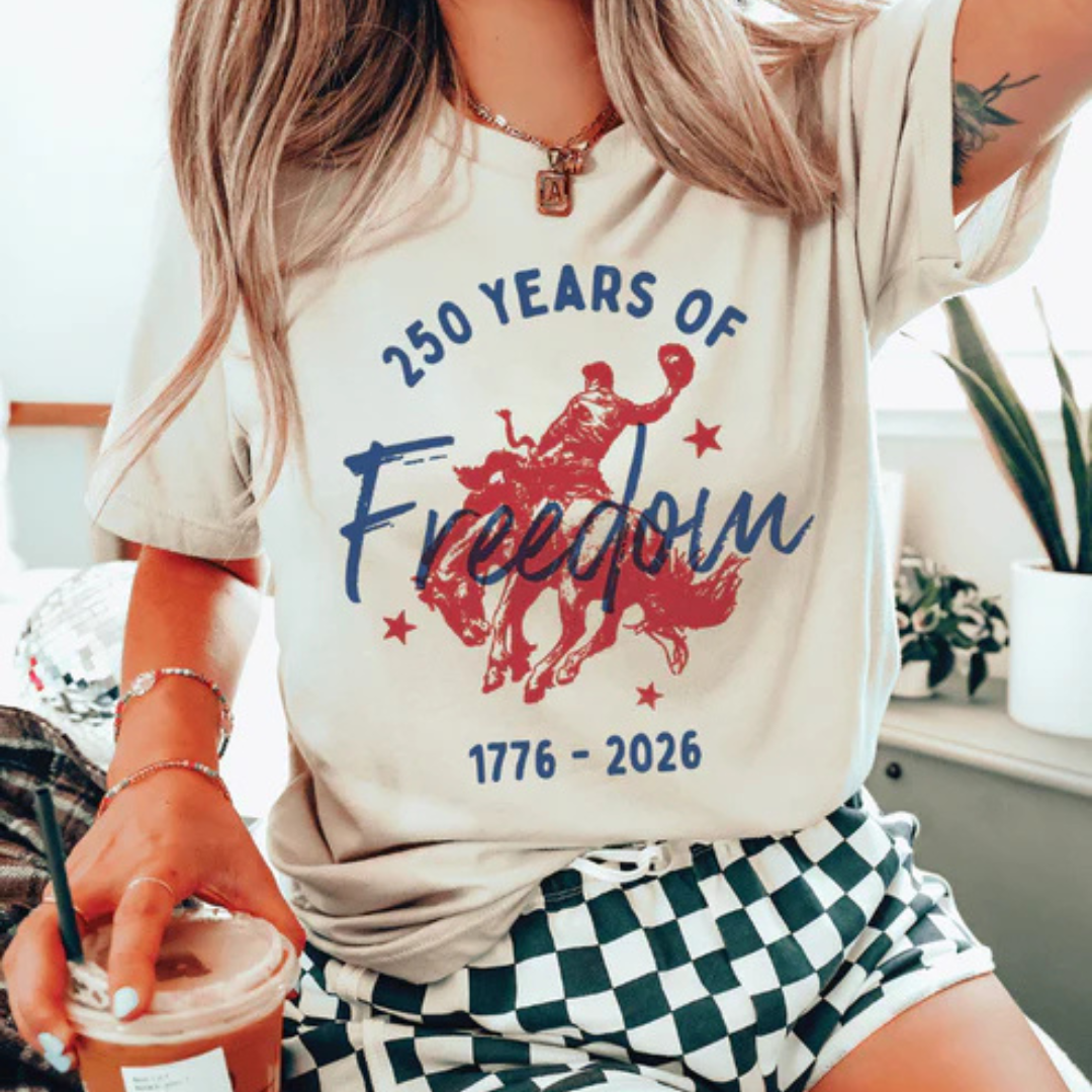 Cream graphic t-shirt featuring a patriotic bronc rider design with 250 Years of Freedom and 1776–2026 commemorative artwork.