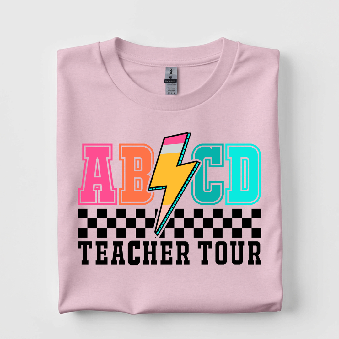Light pink graphic tee featuring a colorful ABCD Teacher Tour design with collegiate lettering, lightning bolt, and black checkered accent.