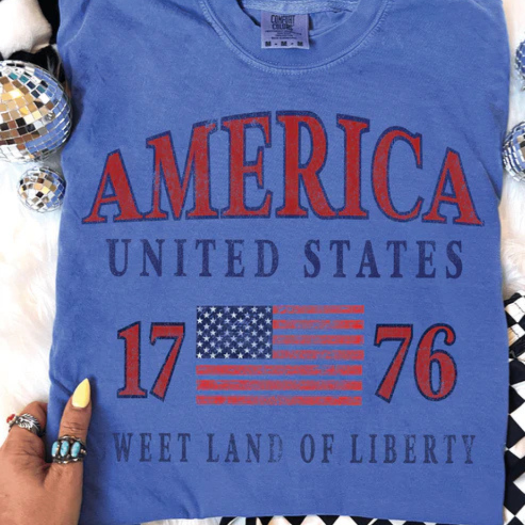Flo Blue Comfort Colors t-shirt featuring a vintage America 1776 patriotic graphic with distressed American flag artwork.