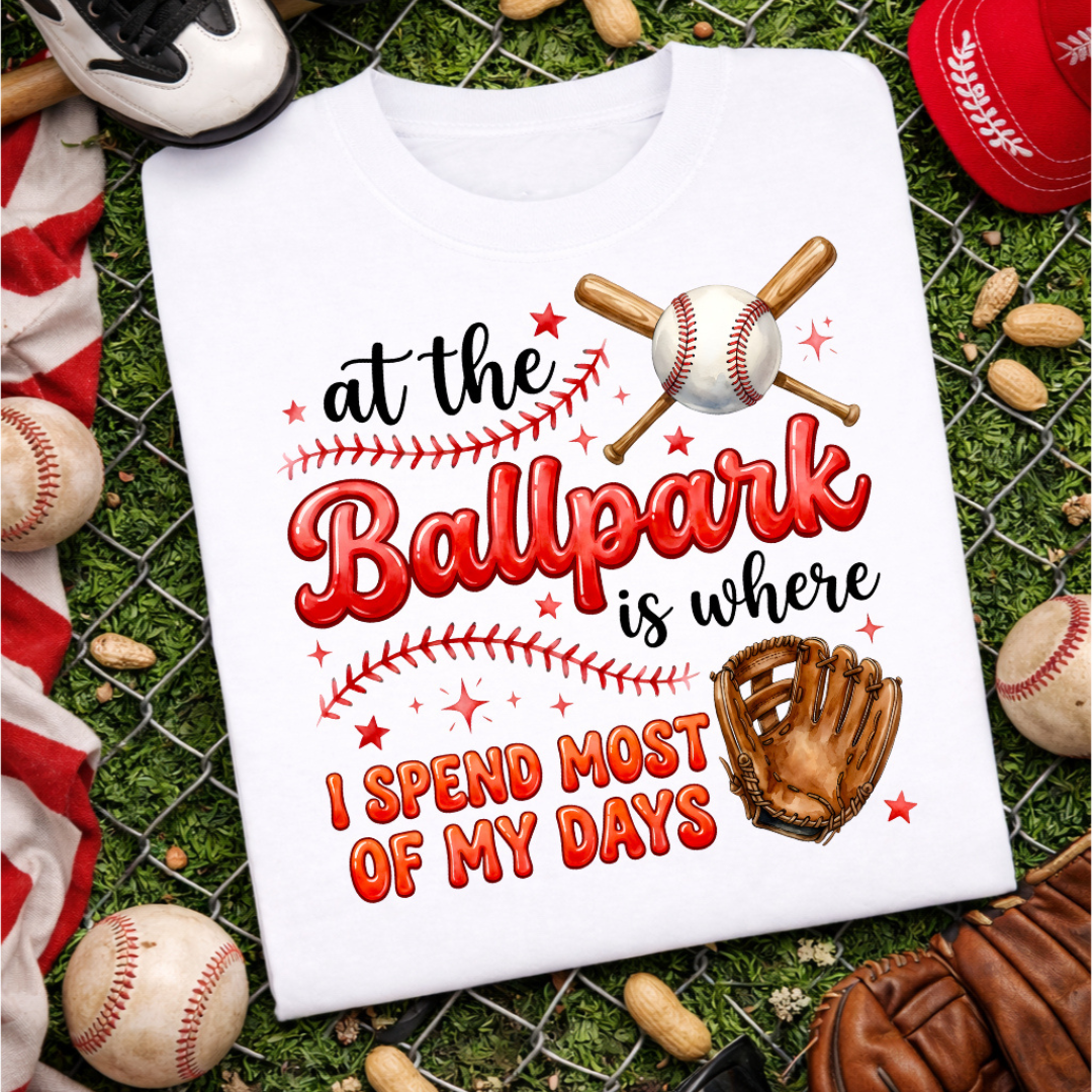 White graphic tee featuring the phrase "At the Ballpark Is Where I Spend Most of My Days" with a baseball, crossed bats, baseball glove, stars, and baseball stitch details.