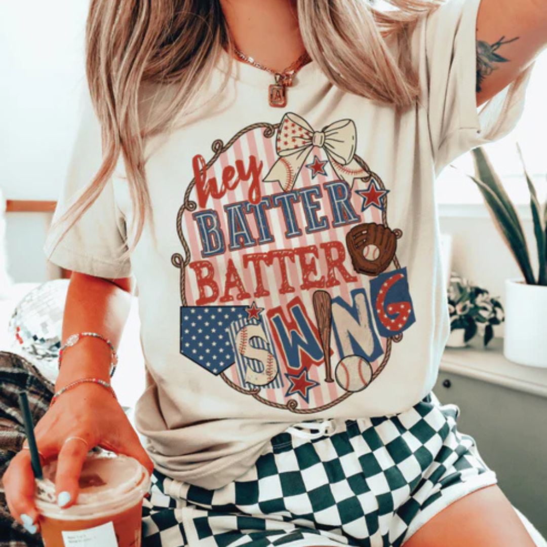 Cream graphic t-shirt featuring a Hey Batter Batter Swing design with baseball-themed artwork, bows, stars, and patriotic details.