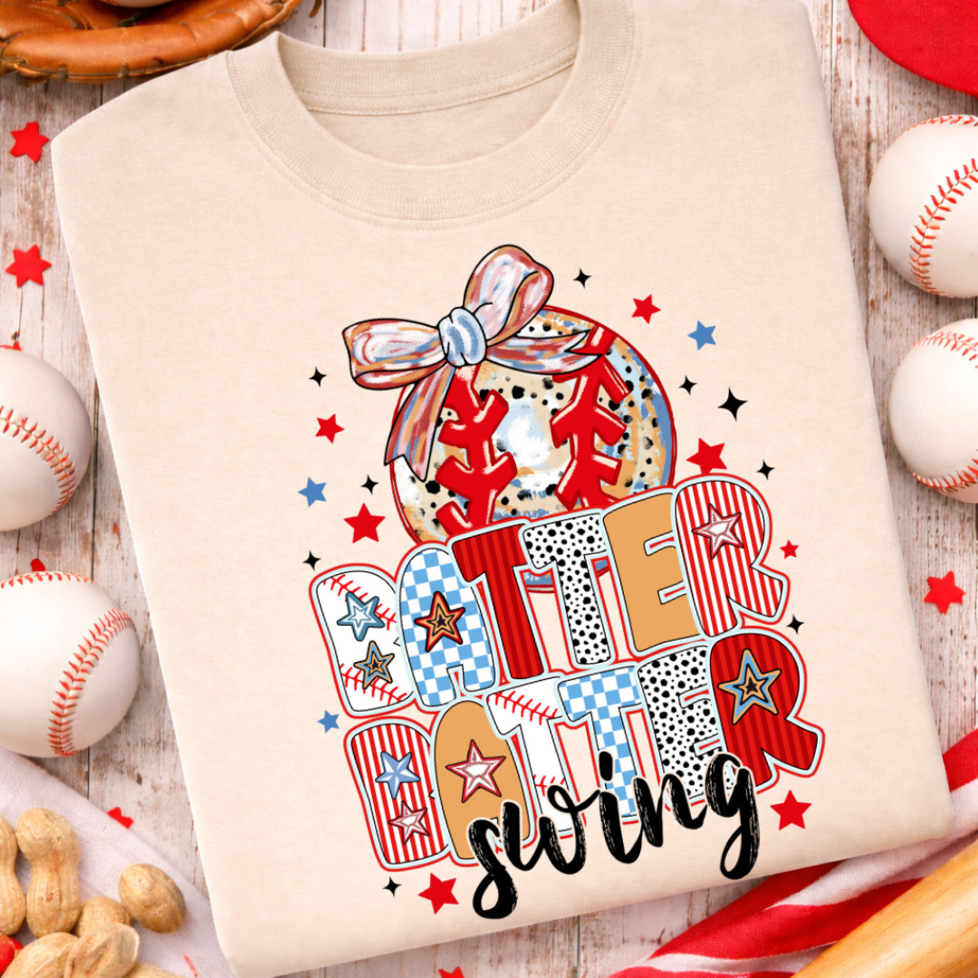 Sand graphic tee featuring colorful "Batter Batter Swing" lettering with a baseball, decorative bow, baseball stitching, and patriotic red, white, and blue accents.