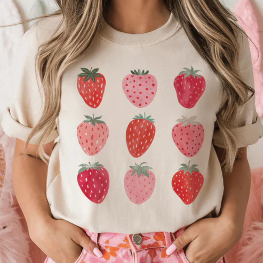Cream graphic tee featuring nine pink and red strawberry illustrations arranged in a grid pattern.