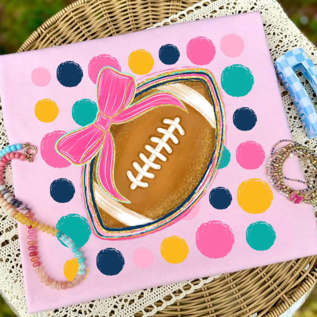 Light pink graphic tee featuring a football with a hot pink bow and colorful polka dots.