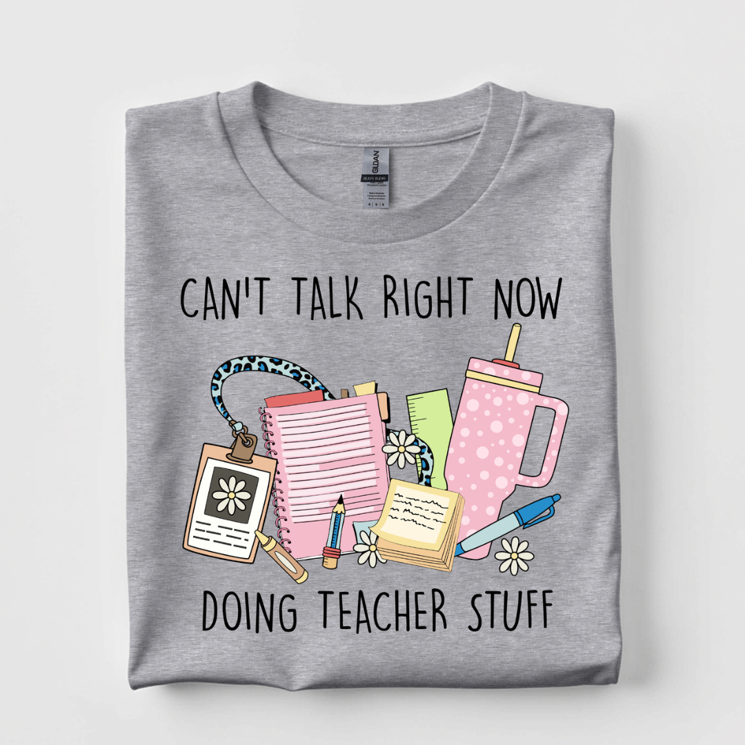 Heather gray graphic tee featuring the phrase "Can't Talk Right Now Doing Teacher Stuff" with colorful classroom supplies, planner, tumbler, and daisy illustrations.