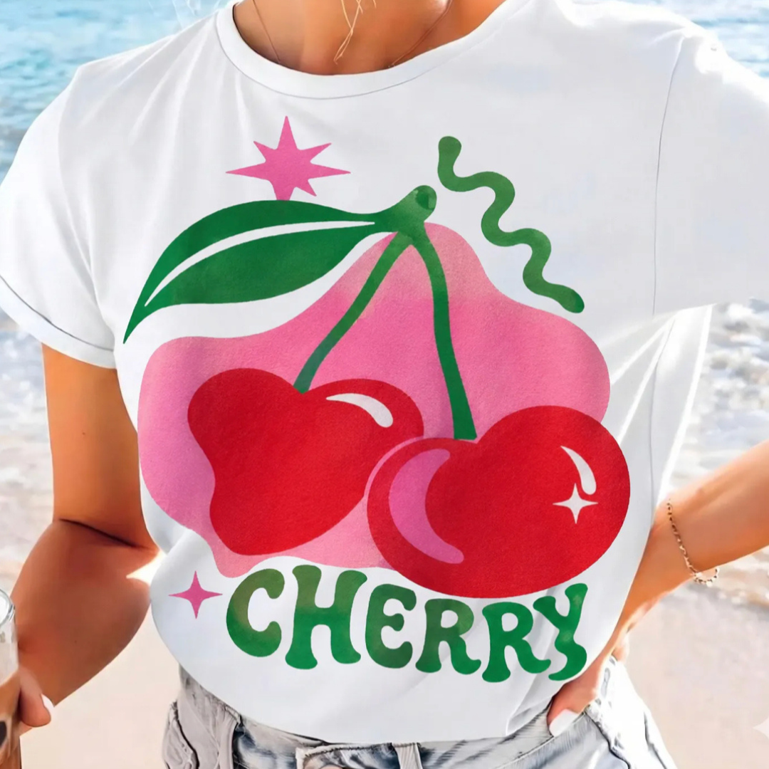 White graphic tee featuring oversized red cherries, pink accents, green lettering, and a retro-inspired cherry graphic design.