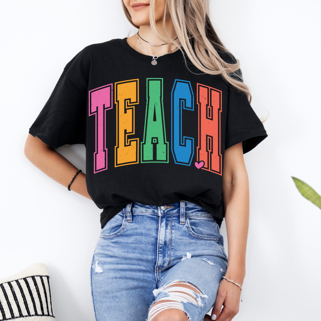 Black graphic tee featuring oversized multicolor varsity-style TEACH lettering with a small heart accent.
