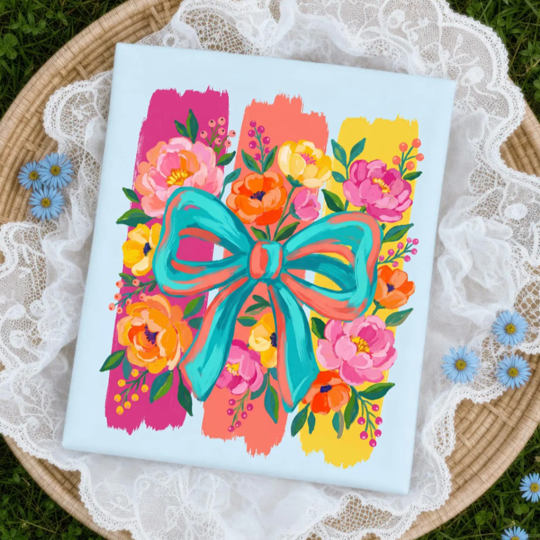 Light blue graphic tee featuring colorful flowers, vibrant brushstroke stripes, and a large turquoise bow centerpiece.